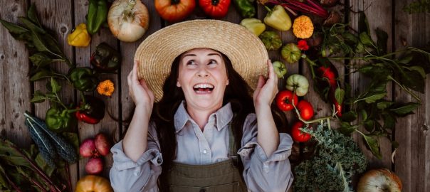 A joyful gardener smiles while lying on a rustic wooden deck, surrounded by an array of colorful vegetables from her garden. Wearing a wide-brimmed hat, her expression conveys the satisfaction of a successful harvest. Perfect for depicting sustainable living and homegrown food.